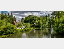 Eine ländliche Szene mit einem kleinen Fluss, umgeben von dichtem Grün und vereinzelten Häusern mit Reetdächern. Im Vordergrund sind Wasserpflanzen und Bäume zu sehen, während die Landschaft von wolkigem Himmel überdeckt wird.