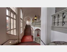 Historisches Treppenhaus mit rotem Bodenbelag, hohen weißen Wänden und großen Rundbogenfenstern auf der linken Seite. Dekorative graue Balustraden und Hängeleuchten prägen das helle, lichtdurchflutete Ambiente.