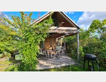 Holzhütte mit Satteldach in einem Garten, umgeben von dichtem Grün. Vor der Hütte befindet sich eine Holzterrasse mit einem runden Tisch, vier Stühlen und einem Grill im Vordergrund.