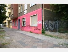 Straßenszene mit einem mehrstöckigen Gebäude in einer Wohngegend; die Fassade ist in Rosa und Braun gestrichen und mit Graffiti bedeckt. Rechts führt ein Gehweg entlang eines Zauns, hinter dem sich überwachsene Vegetation befindet.