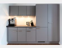 Graue Einbauküche mit Oberschränken, Unterschränken und einer Arbeitsfläche, auf der sich ein Kaffeevollautomat, ein Wasserkocher und ein Messerblock befinden; dezente Beleuchtung unter den Oberschränken.