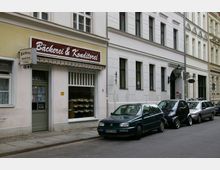 Straßenansicht einer Bäckerei und Konditorei mit gelb-weißer Fassade, großem Schaufenster und Markise mit Schriftzug. Mehrere Autos sind entlang der gepflasterten Straße vor dem Gebäude geparkt.