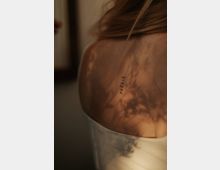 Nahaufnahme eines weiblichen Rückens mit einem kleinen Schriftzug-Tattoo, das das Wort „POÉSIE“ zeigt. Weiches Licht und Schatten von Blättern fallen auf die Haut und erzeugen eine natürliche, warme Atmosphäre.
