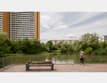 Eine Uferpromenade mit einem kleinen Teich, umgeben von dichter Vegetation und mehrstöckigen Wohngebäuden im Hintergrund. Im Vordergrund steht eine Sitzbank, daneben lehnt eine Person an einem Geländer und schaut zum Wasser.