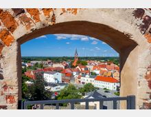 Blick durch einen gemauerten Torbogen auf eine Kleinstadt mit roten und weißen Gebäudedächern, einer zentralen Kirche mit hohem Turm und einer Landschaft mit Windrädern im Hintergrund. Der Himmel ist blau mit wenigen Wolken.