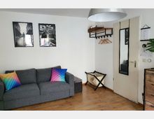 Ein kleines Wohnzimmer mit Holzboden, grauem Sofa und bunten Kissen. An der weißen Wand hängen zwei Schwarz-Weiß-Bilder, daneben befindet sich eine weiße Tür mit einem Spiegel, ein Wandregal mit Kleiderhaken und ein faltbarer Gepäckständer.