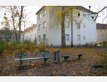 Wohnsiedlung mit einem dreistöckigen Gebäude und hellgrauer Fassade, umgeben von Herbstbäumen, Sträuchern und Laub auf dem Boden; im Vordergrund eine kleine Sitzgruppe mit zwei Holzbänken und einem Mülleimer.