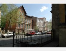 Straßenszene mit historischen Wohngebäuden aus rotem und hell verputztem Backstein, gesäumt von Bäumen. Im Vordergrund ein schmiedeeisernes Metallgitter und Mülltonnen, dahinter geparkte Autos auf einer gepflasterten Straße.