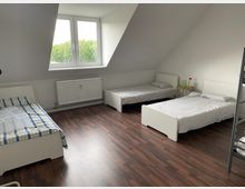 Ein Dachzimmer mit drei Einzelbetten, holzfarbenem Laminatboden und weißen Wänden. Ein großes Fenster mit Blick ins Grüne und ein Heizkörper befinden sich an der Stirnwand; neben einem Bett steht ein kleiner, runder Beistelltisch mit Vase und Blume.