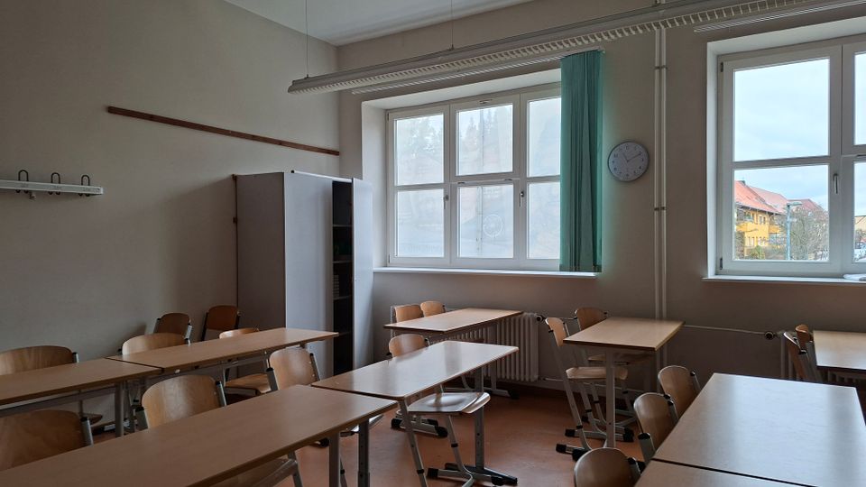Leeres Klassenzimmer mit mehreren Holztischen und Stühlen, einem hohen Schrank und einer Garderobenleiste an der Wand. Große Fenster lassen Tageslicht herein; daneben hängen eine Uhr und ein grüner Vorhang, unter den Fenstern befinden sich Heizkörper.