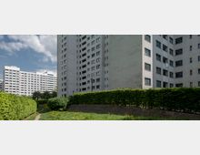 Mehrstöckige Wohngebäude aus Beton mit zahlreichen Fenstern und Balkonen, umgeben von gepflegten Hecken, Wiesen und einem Gehweg in einer Wohnsiedlung. Der Himmel ist teils bewölkt, und vereinzelte Bäume befinden sich im Hintergrund.