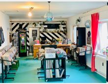 Ein Innenraum eines Schallplattenladens mit türkisfarbenem Boden, mehreren Regalen und Wagen voller Schallplatten. Im Hintergrund ein Tresen mit einer dekorativen Wand in schwarz-weißem Streifenmuster, darüber Pflanzen, Lampen und eine Diskokugel. Links und rechts befinden sich Fenster mit roten Vorhängen und Dekorationsgegenständen.