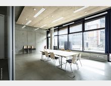 Moderner Besprechungsraum mit großen Fensterfronten, die den Blick nach draußen auf ein Industriegebäude freigeben. Der Raum hat eine Betondecke, helle Tisch- und Stuhlmöbel sowie einen gefliesten Boden.