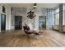 Ein minimalistischer Raum mit Holzboden, weißen Wänden, die mit großflächigen schwarzen Kalligrafien in asiatischen Schriftzeichen bedeckt sind. Drei Türen führen in angrenzende Bereiche, und ein Fenster auf der rechten Seite lässt Tageslicht herein; eine Person liegt entspannt auf einem Ledersessel.