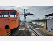 Ein Industriehafen mit einem Schienenfahrzeug auf einem Gleis, das entlang einer Betonpier führt. Im Hintergrund sind ein großer Ladekran, ein Fluss und Bäume am Ufer zu sehen, bei bewölktem Himmel.