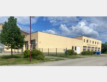 Einstöckiges, hellgelbes Gebäudekomplex mit Flachdach und der Aufschrift "Rettungswache Neuenhagen". Vor dem Gebäude stehen ein Baum, Büsche und ein Zaun, im Hintergrund ist ein teils bewölkter Himmel sichtbar.