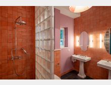 Badezimmer mit roten Wandfliesen und einer Duschkabine, die durch eine Glasbausteinwand abgetrennt ist. Zwei weiße Waschbecken mit ovalen Spiegeln an einer lila gestrichenen Wand, daneben ein Fenster mit Tageslicht.