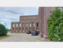 Backsteingebäude mit einfachen geometrischen Strukturen und hohen Fenstern, umgeben von einer gepflasterten Einfahrt mit parkenden Autos. Links ist eine grüne Hecke und eine Laterne, rechts befinden sich weitere Backsteinfassaden und ein Busch.