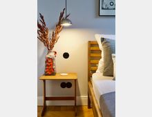 Nachttisch aus Holz neben einem Bett mit Holzkopfteil, darauf eine Vase mit Trockenblumen und eine kleine Lampe, die an der Wand hängt und warmes Licht abstrahlt; darunter befinden sich zwei Steckdosen. Im Hintergrund eine weiße Wand mit einem gerahmten Bild und Parkettboden.