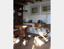 Ein gemütlicher, traditionell eingerichteter Raum mit Holzmöbeln, darunter ein runder Tisch mit zwei unterschiedlichen Stühlen. An der Wand hängen Gemälde und Regale, die mit Büchern, Statuen und kleinen Objekten dekoriert sind.