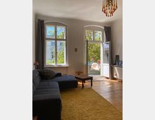 Wohnzimmer mit einer großen grauen Eckcouch, einem gelben Teppich und Holzboden. Im Hintergrund befinden sich zwei hohe Fenster und eine geöffnete Tür, die auf einen Balkon mit Pflanzenmöbeln und Grünblick führt.