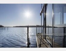 Moderne gläserne Gebäudestruktur an einem Seeufer mit Holzsteg, reflektierendem Wasser und einer hellen Sonne am klaren blauen Himmel. Im Hintergrund sind bewaldete Uferlinien zu erkennen.