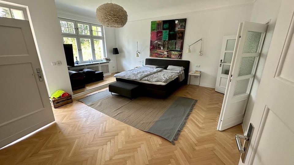 Ein helles Schlafzimmer mit Fischgräten-Holzfußboden, einem Doppelbett mit dunklem Gestell, einer modernen Hängelampe und einem großen Fenster, das viel Tageslicht hereinlässt. An der Wand hängt ein buntes Gemälde, und daneben stehen Nachttische mit Leselampen.