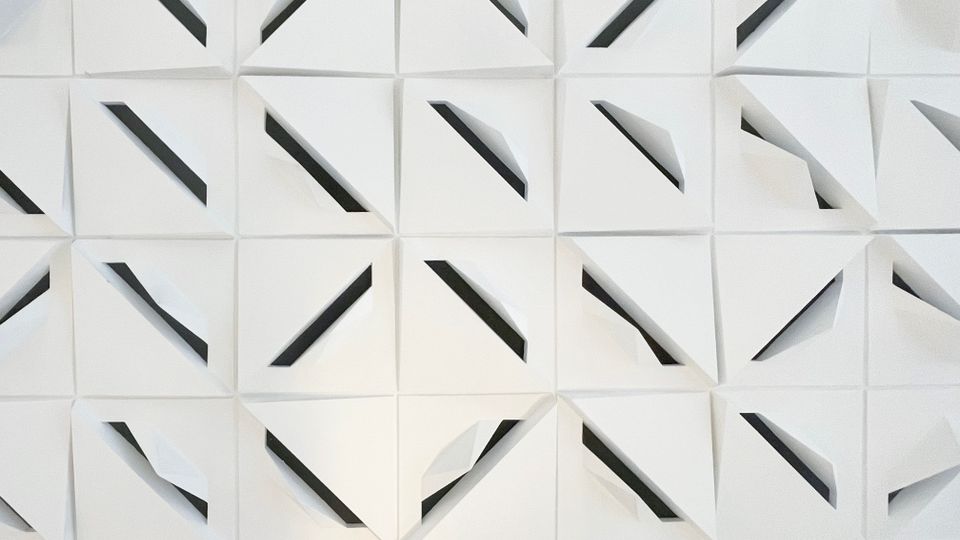 Nahaufnahme einer modernen, weißen Wand- oder Deckenverkleidung aus quadratischen Paneelen mit gefalteten, dreidimensionalen Dreiecksformen. Zwischen den gefalteten Flächen verlaufen diagonal angeordnete dunkle Schlitze, die ein geometrisches Muster bilden.