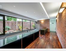 Moderner Innenraum mit roten Backsteinwänden, Holzfußboden und einer großen Fensterfront, die zu einem Außenbereich mit Holztischen und -stühlen sowie einer Ziegelmauer führt. Der Raum enthält eine Glasvitrine, eine Sitzbank und eine Tür zu sanitären Einrichtungen.