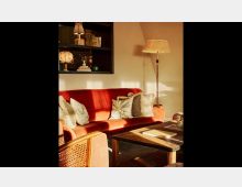 Wohnzimmer mit einem roten Samtsofa, dekoriert mit mehreren Kissen, einer Stehlampe mit hellem Schirm und einem Holztisch im Vordergrund. Im Hintergrund ein Regal mit Büchern und dekorativen Objekten.