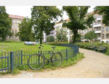 Ein kleiner städtischer Park mit einer Wiese, umgeben von einem niedrigen Metallzaun, auf dem ein Fahrrad lehnt. Im Hintergrund stehen mehrstöckige Wohnhäuser mit Balkonen sowie Bänke entlang eines Kieswegs; mehrere Bäume spenden Schatten.