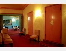 Flur eines Theaters oder Konzerthauses mit roten Teppichböden und gelben Wänden. Auf der rechten Seite befinden sich nummerierte Türen mit der Aufschrift "Loge", links sind gepolsterte Stühle aufgereiht, und im Hintergrund ist ein kleiner Speisebereich mit gedeckten Tischen und einer Bar zu sehen.