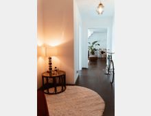 Wohnungsflur mit dunklem Holzfußboden und weiß gestrichenen Wänden. Im Vordergrund ein runder beigefarbener Teppich, ein kleiner Beistelltisch mit Lampe, sowie ein Fahrrad an der Wand; im Hintergrund ein Wohnbereich mit Pflanzen und Esstisch erkennbar.