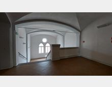 Innenansicht eines Gebäudes mit weiß verputzten Wänden, einem Rundbogen an der Decke und einem Treppenabgang. Im Hintergrund befinden sich drei bogenförmige Fenster mit einem runden Fenster darüber, die Tageslicht in den Raum lassen.