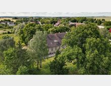 Luftaufnahme eines ländlichen Gebäudes mit Ziegeldach und zahlreichen Fenstern, umgeben von hohen Bäumen und Grünflächen. Im Hintergrund sind eine kleine Siedlung mit Häusern und weite Felder sichtbar.