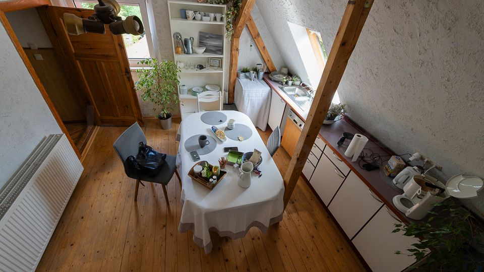 Kleine Dachgeschossküche mit Holzfußboden, sichtbaren Holzbalken und schrägen Wänden; in der Mitte steht ein ovaler Esstisch mit Tischdecke und Stühlen. Entlang der Wand verläuft eine Küchenzeile mit Spüle unter einem Dachfenster, daneben ein offenes Regal mit Geschirr und ein Fenster zur Seite.