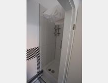 Kleine Duschkabine mit weiß gefliesten Wänden und schwarzem Schachbrettmuster an der unteren Hälfte. Die Ausstattung umfasst eine Handbrause und eine Edelstahlablage an der Wand.