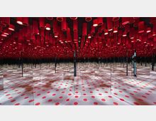 Raum mit verspiegelten Wänden, die eine scheinbar unendliche Reflexion schaffen. Die Decke ist mit zahlreichen roten Zylindern dekoriert, während der Boden mit weißen Flächen und roten Punkten gestaltet ist.