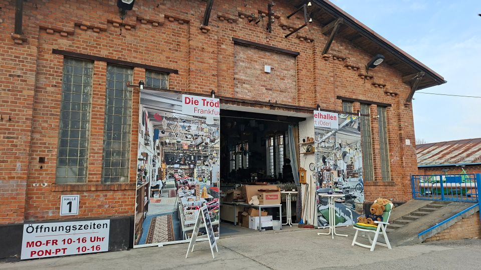 Backsteinfassade einer ehemaligen Industriehalle mit hohen Glasbausteinfenstern und großer, teils geöffneter Toröffnung, an der Werbebanner für einen Trödelmarkt hängen. Vor dem Eingang stehen Aufsteller und einzelne Möbelstücke; rechts führt eine Rampe mit blauem Geländer entlang des Gebäudes.
