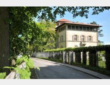 Herrenhaus mit heller Fassade, Fensterläden und dunkelrotem Dach, umgeben von einem eisernen Zaun und dichten Hecken; links ist eine von Bäumen gesäumte Straße sichtbar.