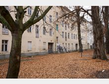 Ein mehrstöckiges, helles Wohngebäude mit sichtbarer Alterung und geschlossenen Fensterläden steht in einer von Bäumen gesäumten Hofanlage, bedeckt von einer Schicht aus Herbstlaub. Die kahle Baumstruktur und die Gebäudefassade weisen auf eine ruhige, herbstliche Atmosphäre hin.