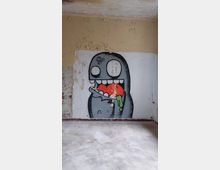 Ein leerer Raum mit abblätterndem Putz an den Wänden. Auf der weißen Wand befindet sich ein farbiges Graffiti eines stilisierten, grauen Charakters, der ein rotes Herz isst.