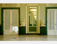 Eleganter Innenraum mit grünen Marmorelementen, zwei weißen Flügeltüren mit Vorhängen, einem zentralen großen Spiegel, der mit einem Kronleuchter reflektiert wird, sowie zwei klassischen weißen Statuen auf Sockeln. In der Mitte steht ein runder Stehtisch mit weißer Tischdecke.