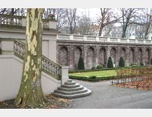 Steingartenanlage mit Treppenaufgang und Balustrade im Vordergrund, dahinter eine lange Mauer mit Rundbögen, die von kahlen Ranken bewachsen ist, umgeben von gepflegten Grünflächen, Büschen und kahlen Bäumen.