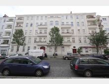 Mehrstöckiges Wohngebäude in einer städtischen Straße mit heller Fassade, Balkonen und einigen Bäumen vor dem Gebäude. Geparkte Autos und ein Kleintransporter stehen am Straßenrand auf Kopfsteinpflaster.