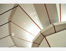 Decke mit geometrischem Design aus weißen, segmentierten Platten, die durch rote und schwarze Linien voneinander getrennt sind; weiche Beleuchtung strahlt zwischen den Platten hervor.