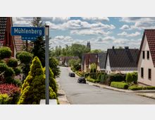 Wohnstraße in einer Vorstadt mit Einfamilienhäusern, die überwiegend rote und graue Dächer haben. Im Vordergrund ist ein Straßenschild mit der Aufschrift „Mühlenberg“ zu sehen, umgeben von gepflegten Hecken und Bäumen; im Hintergrund ist ein Kirchturm erkennbar.