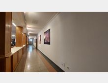 Ein langer, schlichter Flur mit weißen Wänden und einer grauen, mit roten Akzenten durchzogenen Fliesenfläche. An der rechten Wand hängen gerahmte Bilder, während die linke Seite durch Holzpaneele und eine Thekenstruktur geprägt ist.