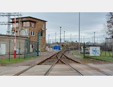 Gleisanlage mit mehreren Eisenbahngleisen, die sich verzweigen, im Vordergrund ein Bahnübergang. Am linken Rand steht ein mehrstöckiges Gebäude aus roten Klinkersteinen, im Hintergrund sind Oberleitungsmasten und Güterwaggons zu sehen.