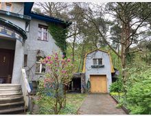 Graues Wohnhaus mit Treppenaufgang und Holzhaustür, daneben ein separates kleines Gebäudeteil mit gelber Holztür und Satteldach. Im Vordergrund steht ein blühender Magnolienstrauch, umgeben von Bäumen und dichter Vegetation.